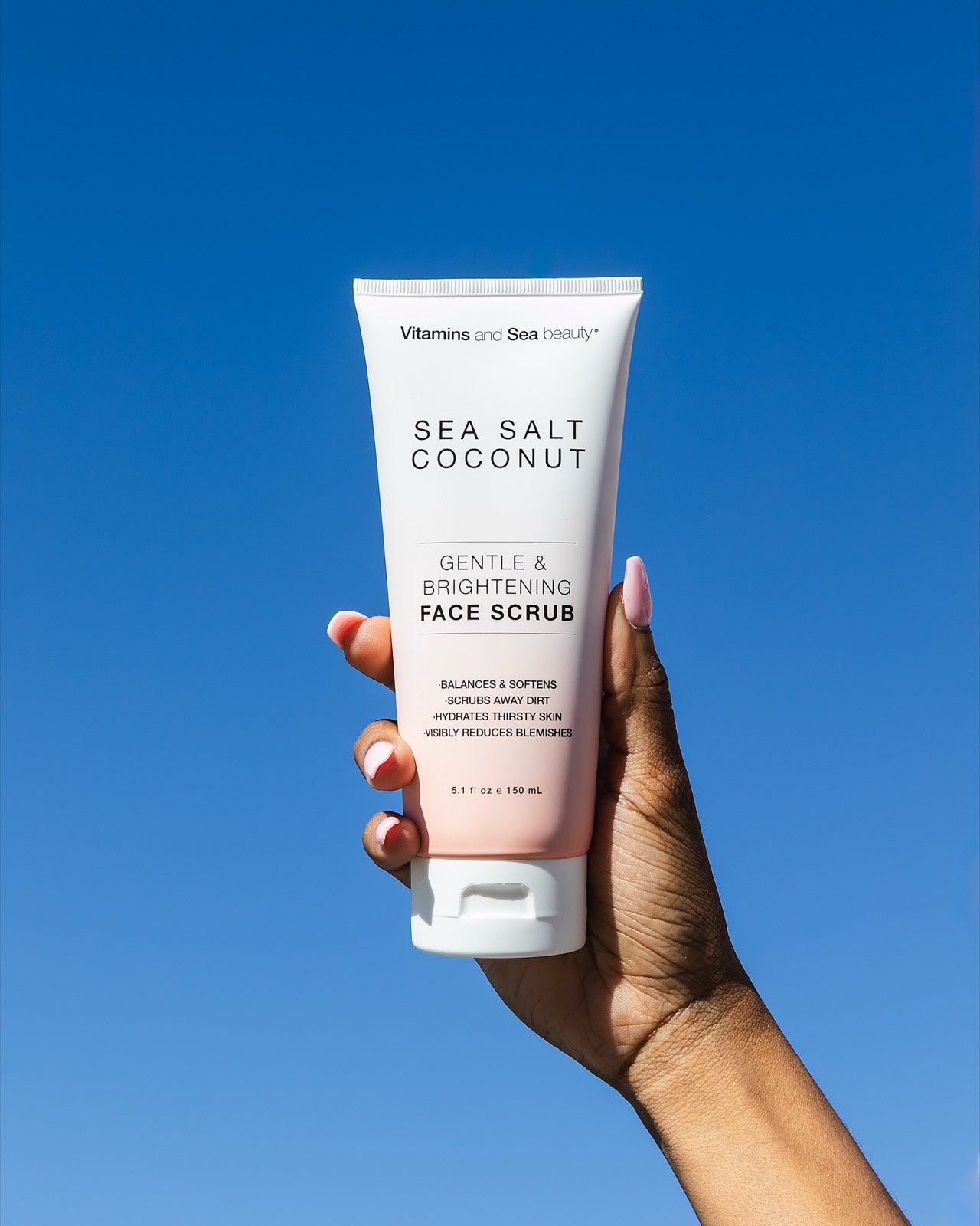 A hand holding the Sea Salt + Coconut face scrub against a deep blue sky